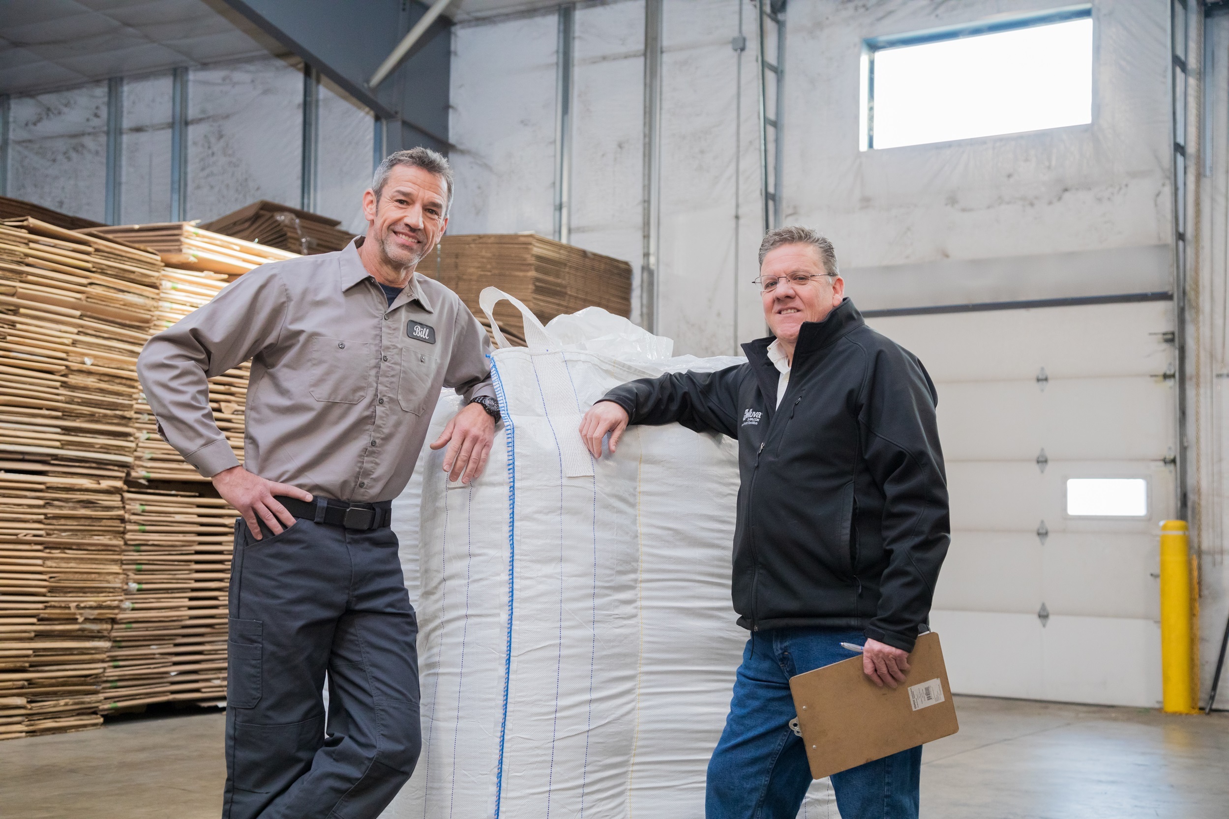 Two men standing in a warehouse next to a bulk bag and smiling at the camera